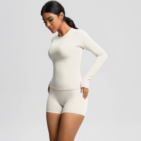 Woman in cream long-sleeve top and shorts, standing sideways against a light gray background.